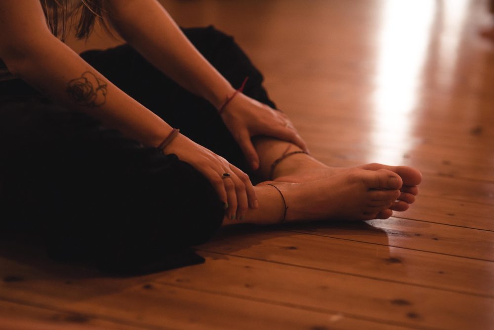 Eine Person sitzt entspannt auf dem Holzboden, die Hände ruhen an den Füßen